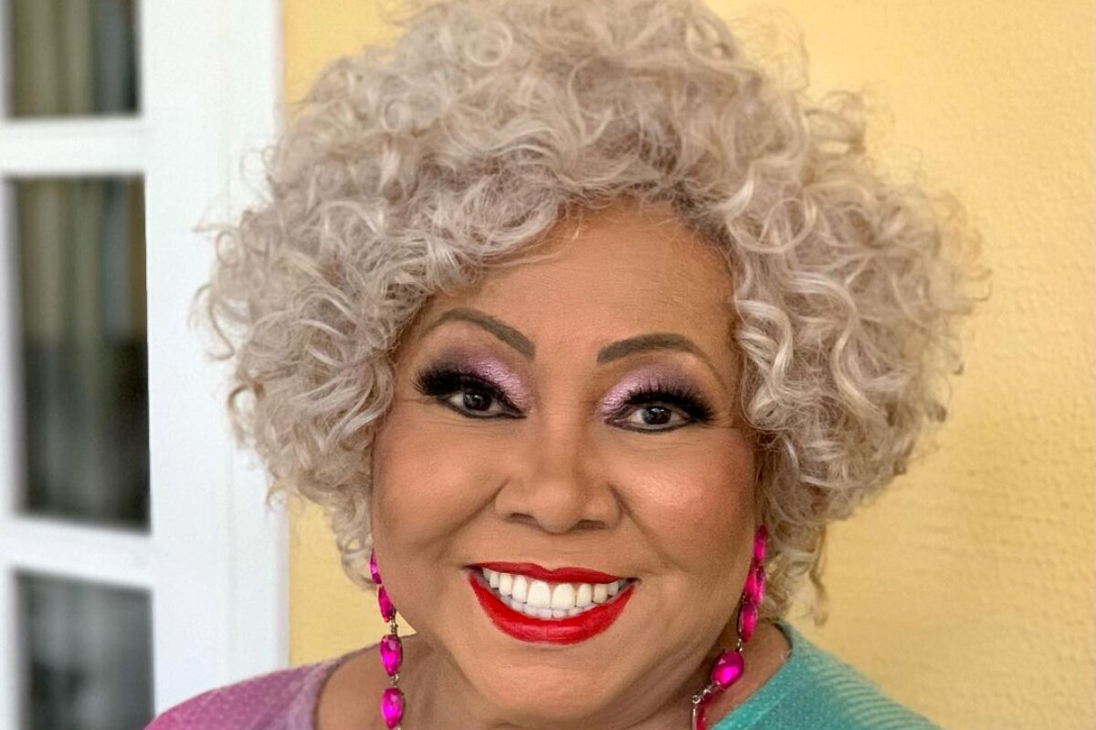 Alcione sorrindo, de cabelos grisalhos, brincos rosa, camisa verde e rosa