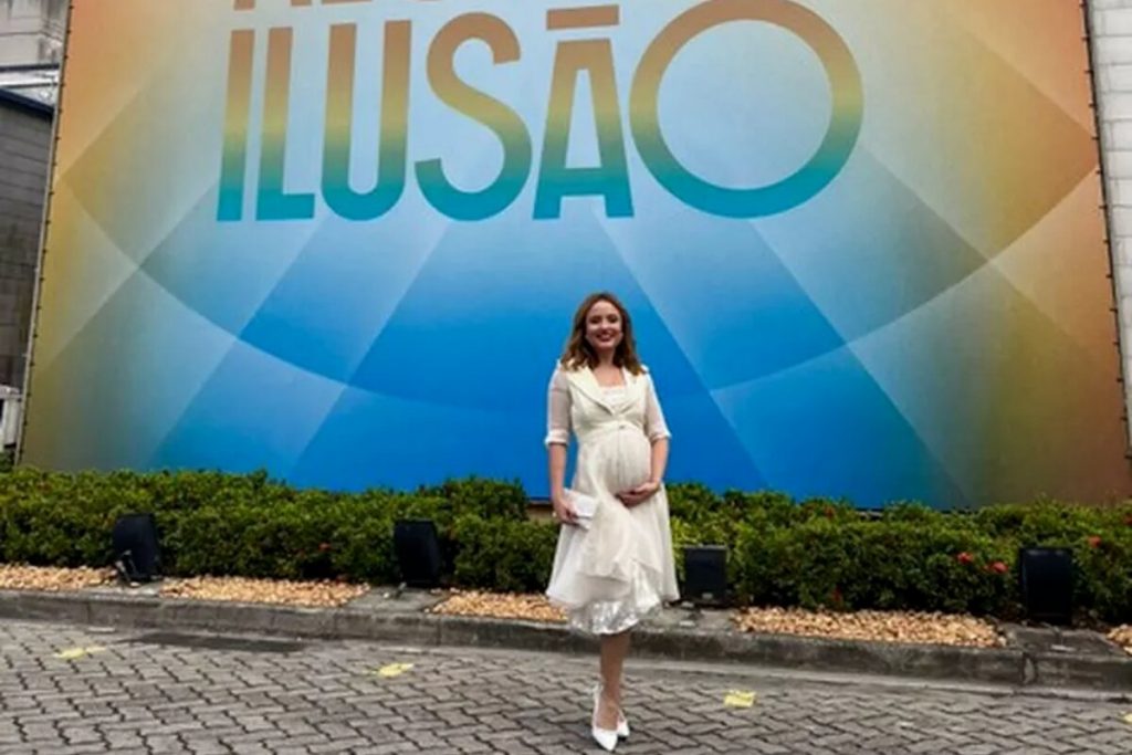 Larissa Manoela de vestido amarelo claro, com barriga cenográfica, na fachada do estúdio de Além da Ilusão