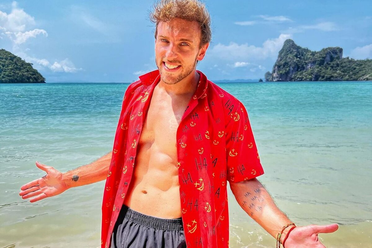 Leo Lins de camisa vermelha aberta na frente, na praia, de braços abertos, sorrindo