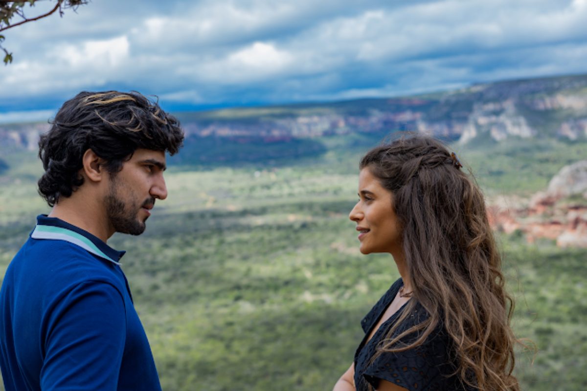 Renato Góes de casaco azul e Isadora Cruz de camisa preta na fictícia Canta Pedra, de 'Mar do Sertão - paisagem verde ao fundo, céu entre nuvens