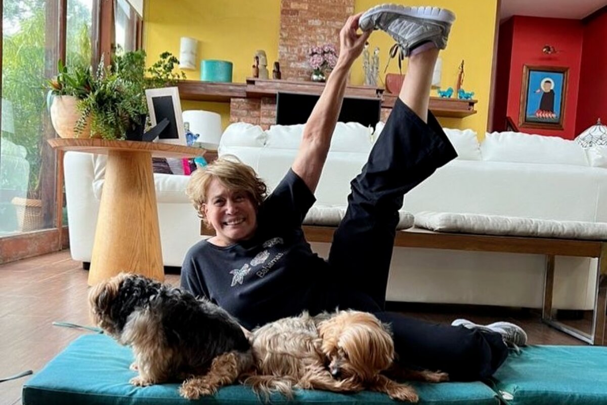 Susana Vieira deitada delado no chão, ao lado dos cachorros, com a perna esquerda esticada para cima, de calça e camisa preta