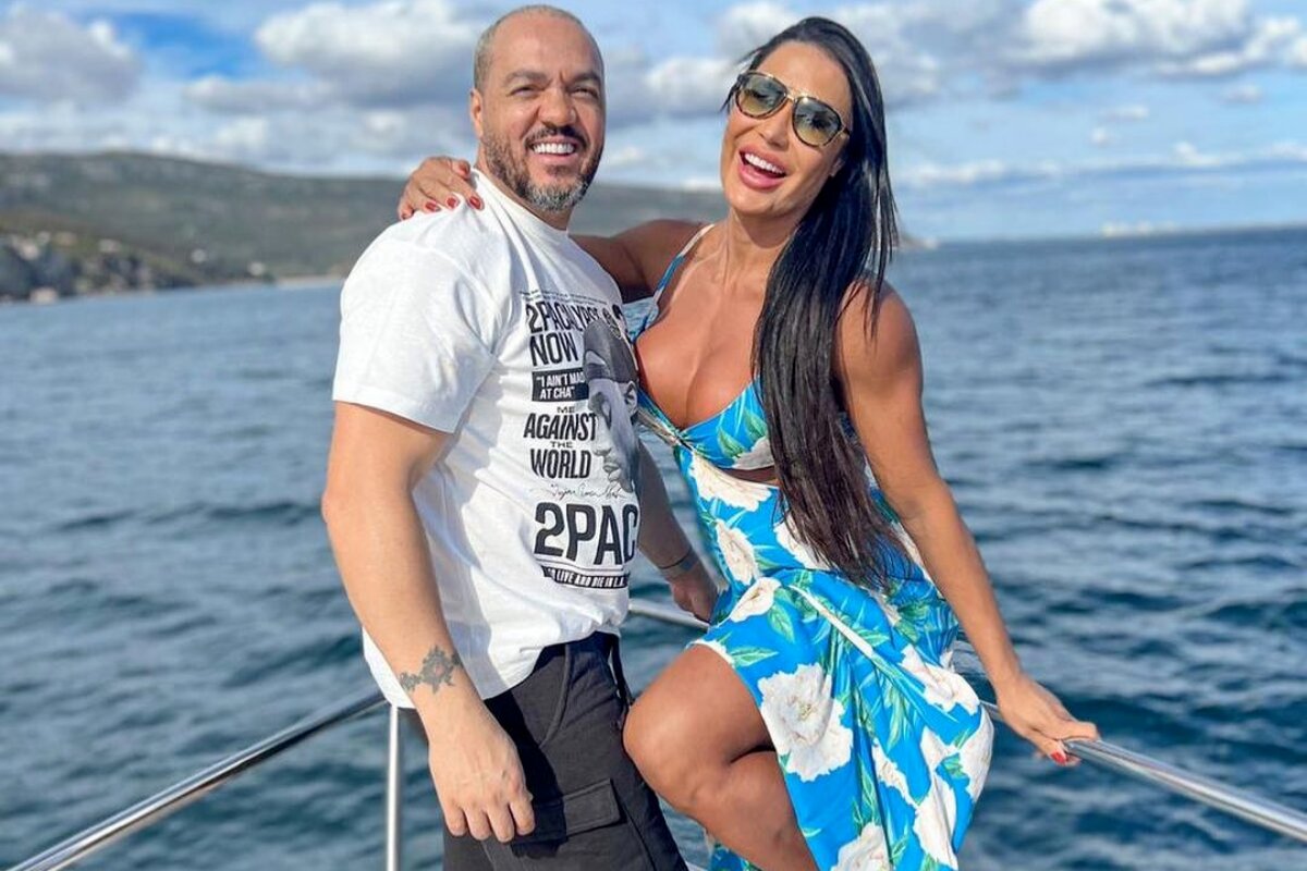 Belo de camisa branca e bermuda preta, com Gracyanne, de óculos escuros e vestido azul e branco, ambos na proa de um barco