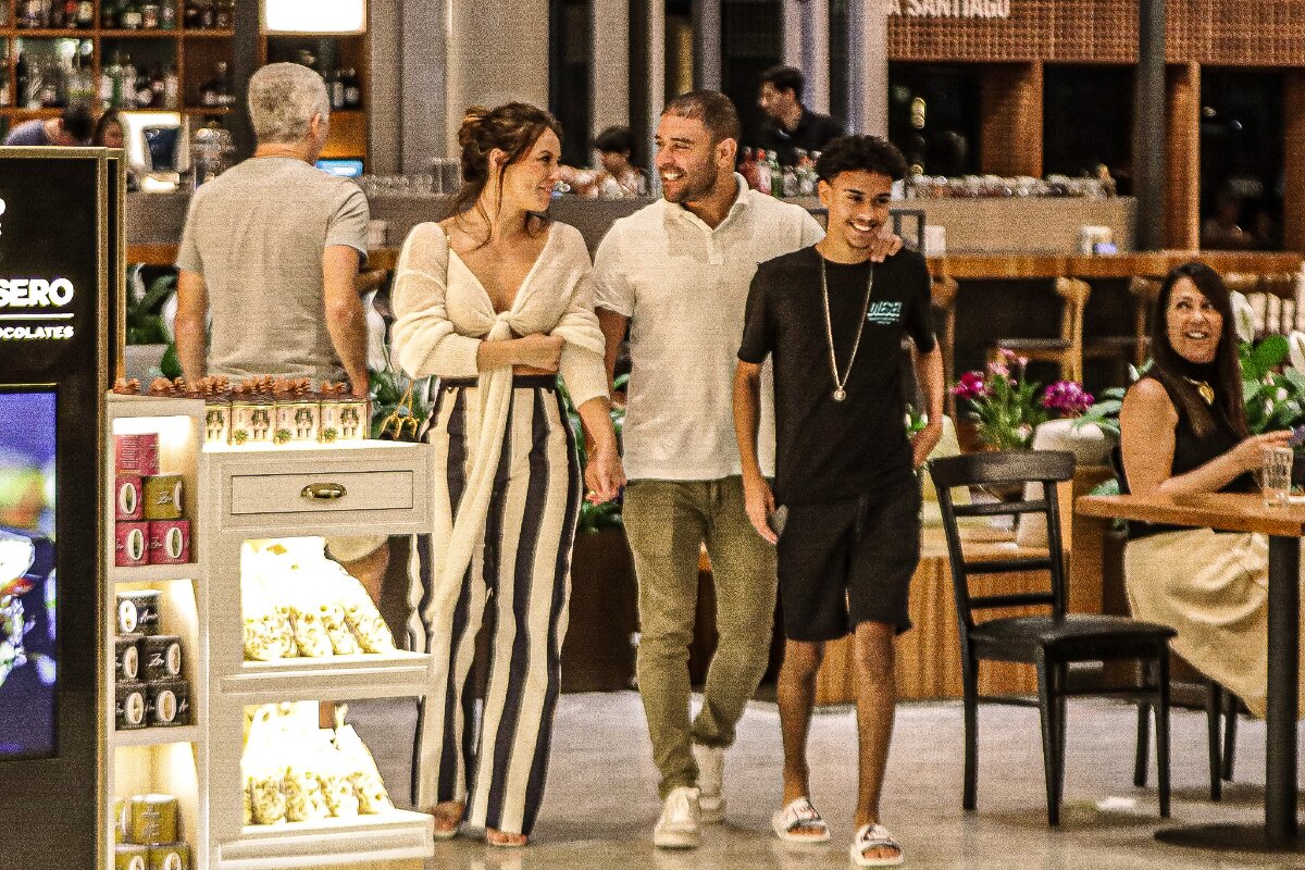 Diogo Nogueira de mãos dadas com Paolla Oliveira, andando no corredor do shopping, ela de calça de listras pretas e brancas, ele de calça marrom e camisa branca, , o cantor abraçado ao filho, Davi, que veste bermuda e camisa preta