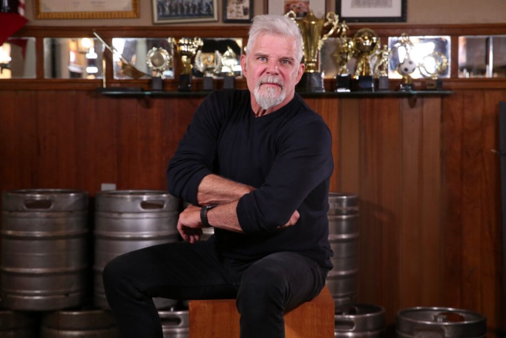 Raul Gazolla de calça e camisa preta, sentado, de braços cruzados, no cenário do bar de Travessia