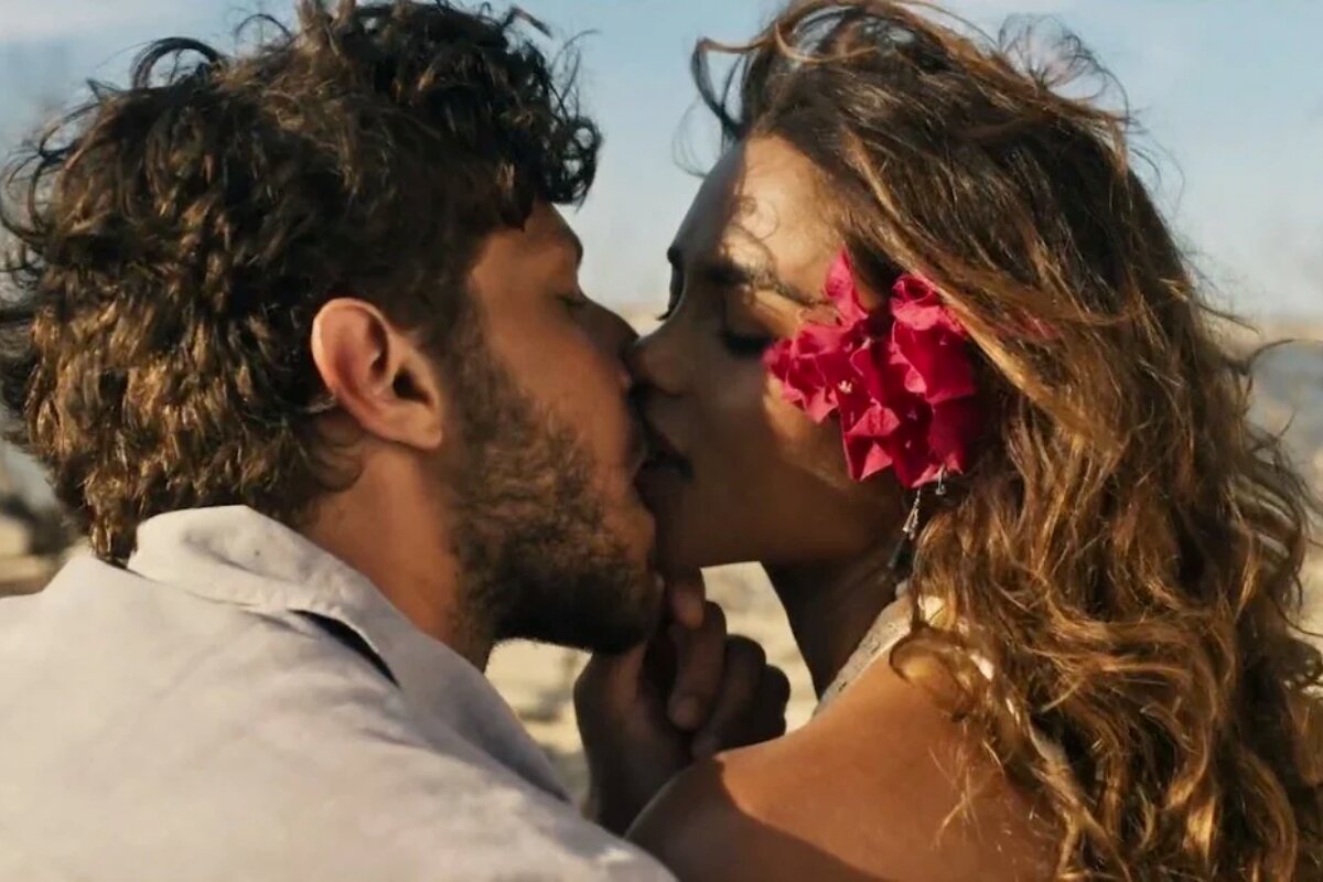 Chay Suede e Lucy Alves caraterizados como Ari e Brisa, ela com uma flor vermelha no cabelo, ambos sentados, se beijando