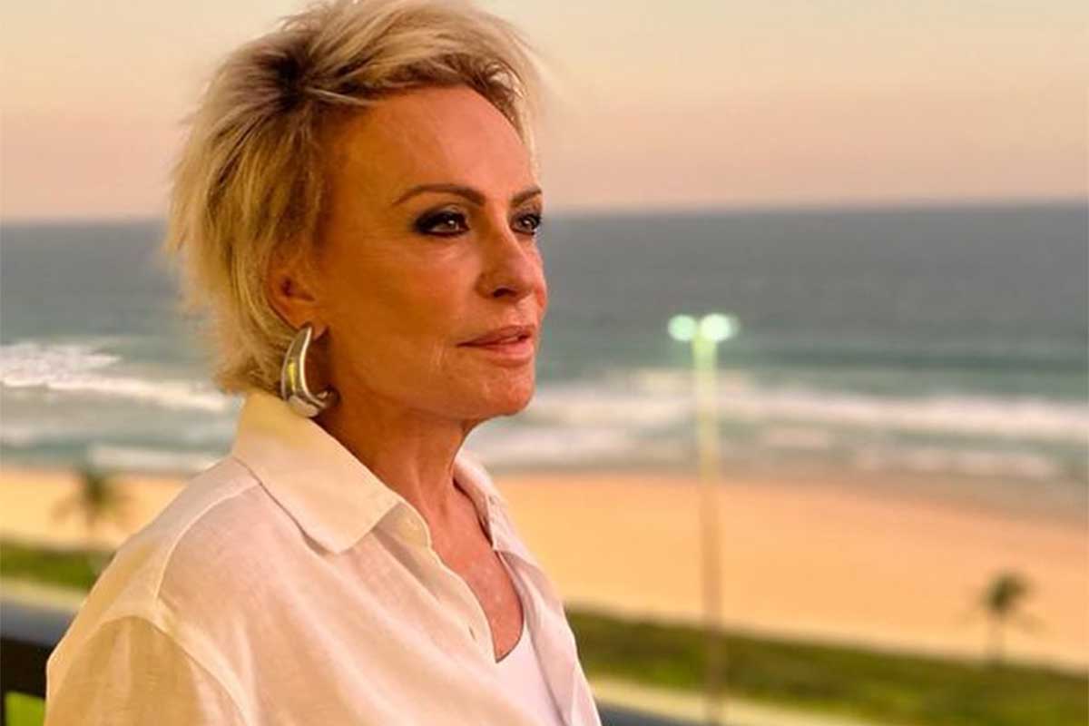 Ana Maria Braga, de camisa branca, na varanda, olhando o por do sol na praia