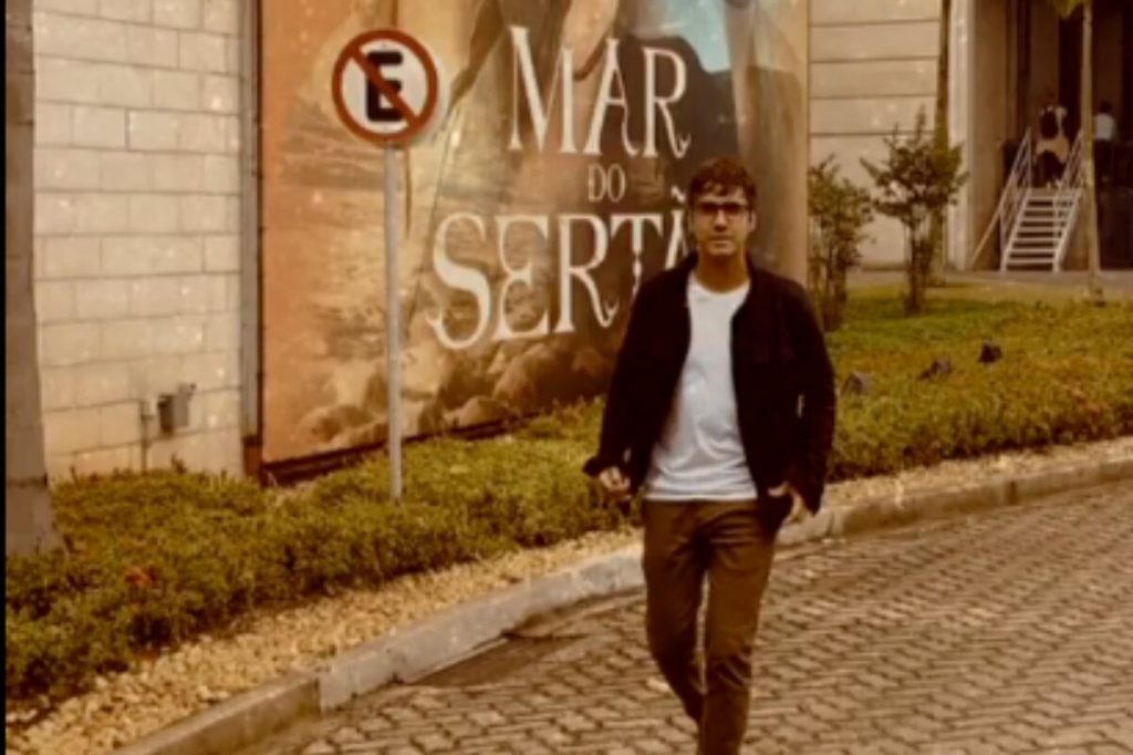 Gabriel Godoy andando de calça marrom, camisa branca e casaco preto, em frente ao estúdio de "Mar do Sertão"