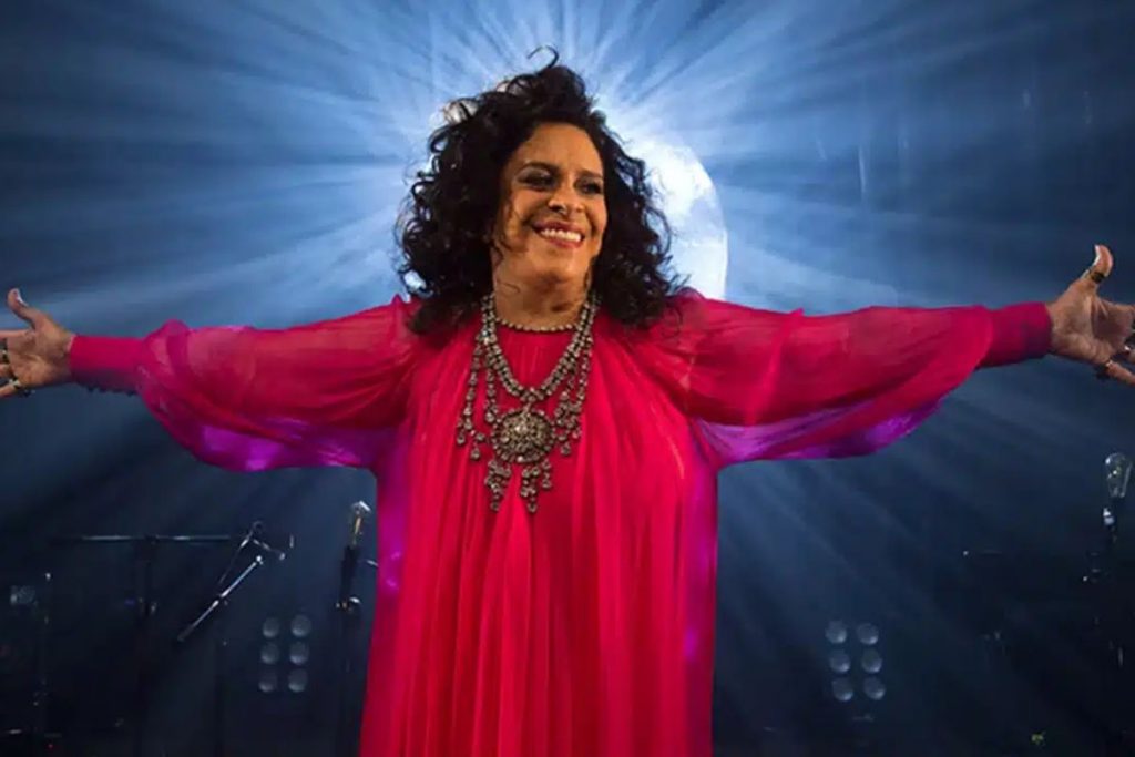 Gal Costa de vestido vermelho, com os braços abertos e iluminação de fundo