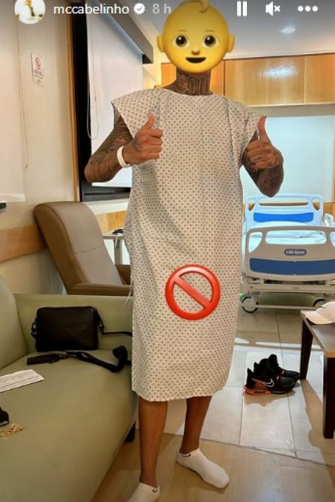 MC Cabelinho com roupa de internação, fazendo sinal de positivo, em pé, no hospital