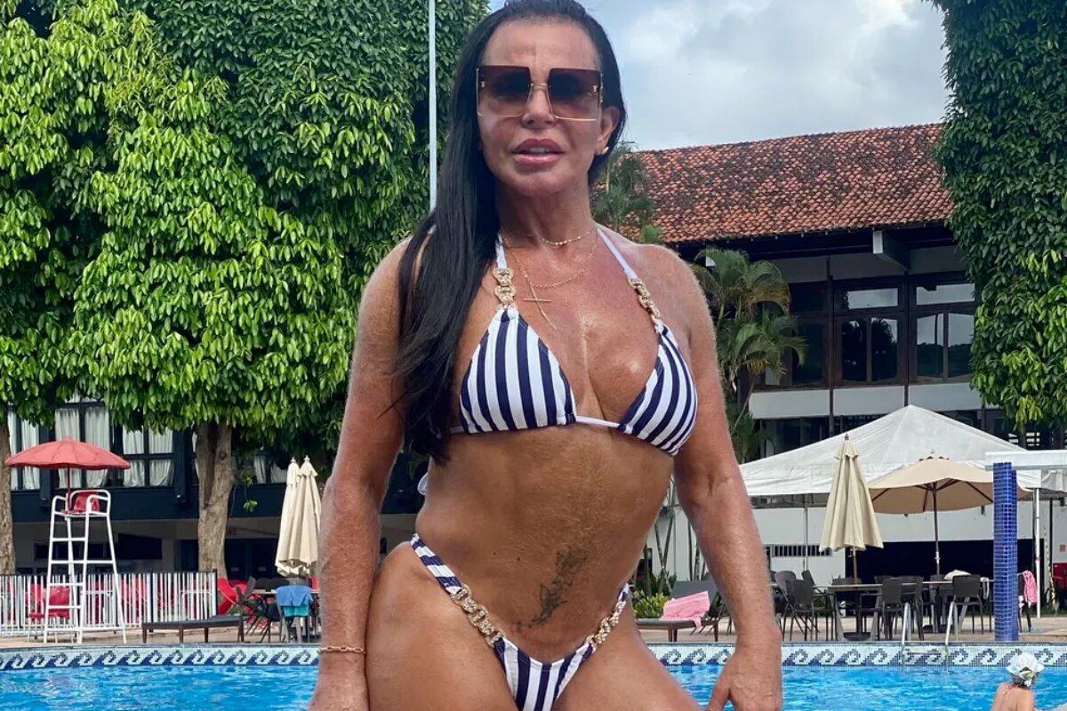 Gretchen de biquíni azul e branco de listras e óculos escuros, com uma piscina ao fundo
