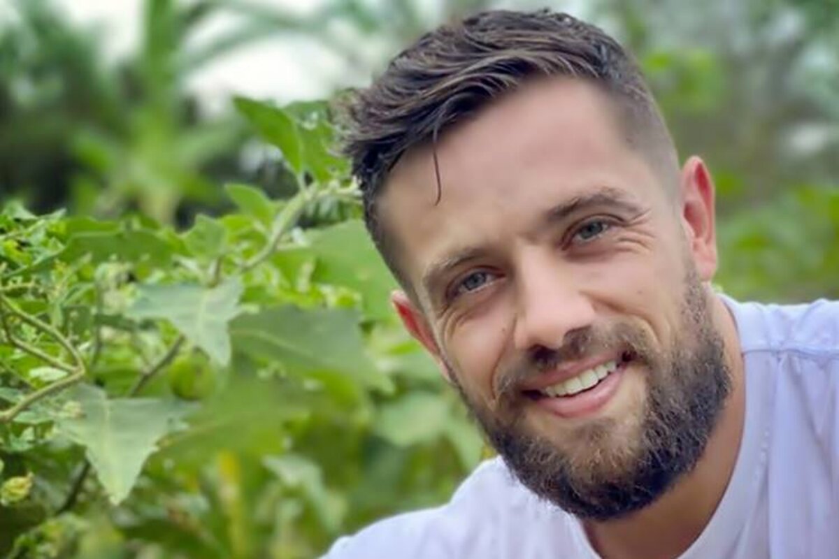 Close de Rafael Cardoso sorrindo, de camisa branca, em uma plantação