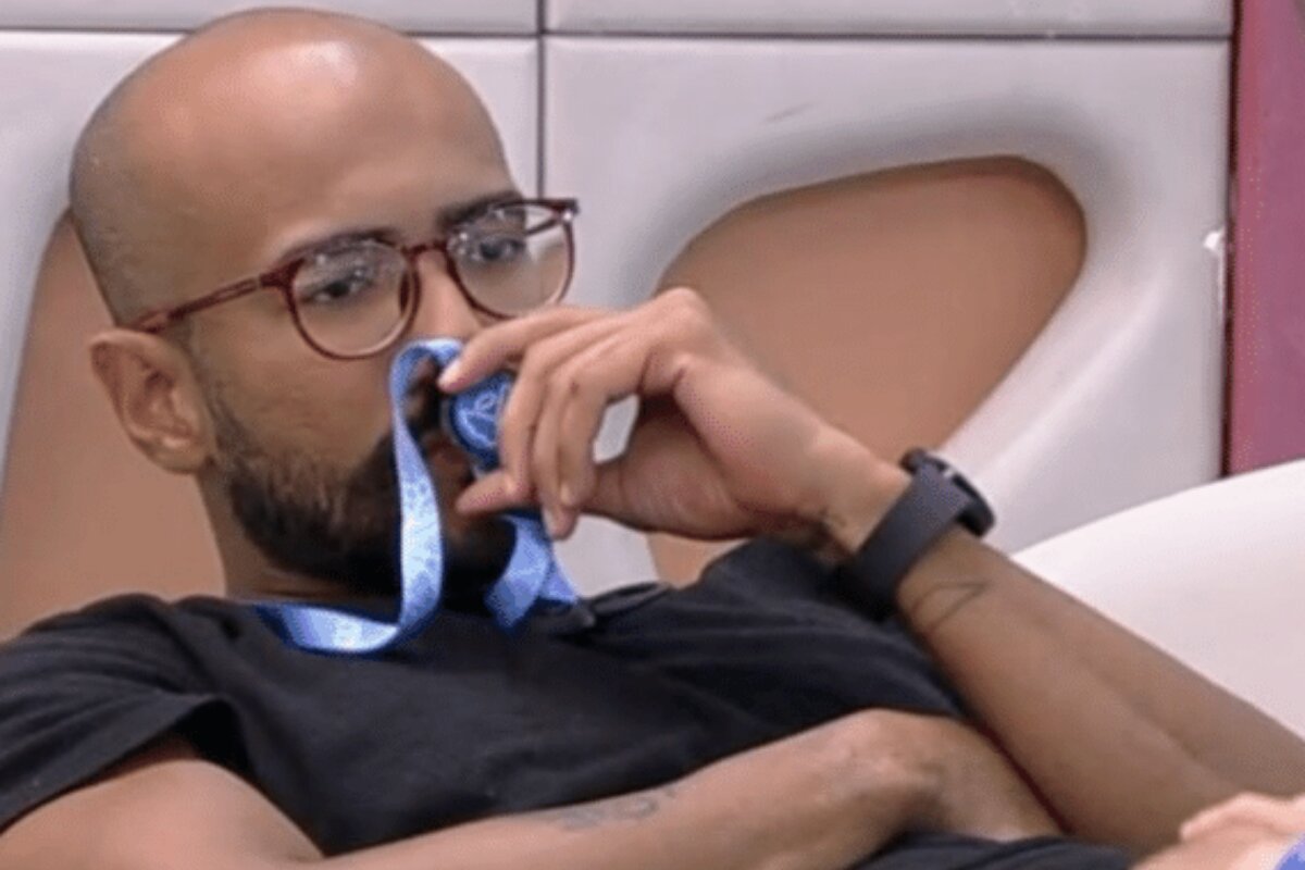 BBB 23 - Ricardo deitado, com o colar do anjo no pescoço, medalha na boca