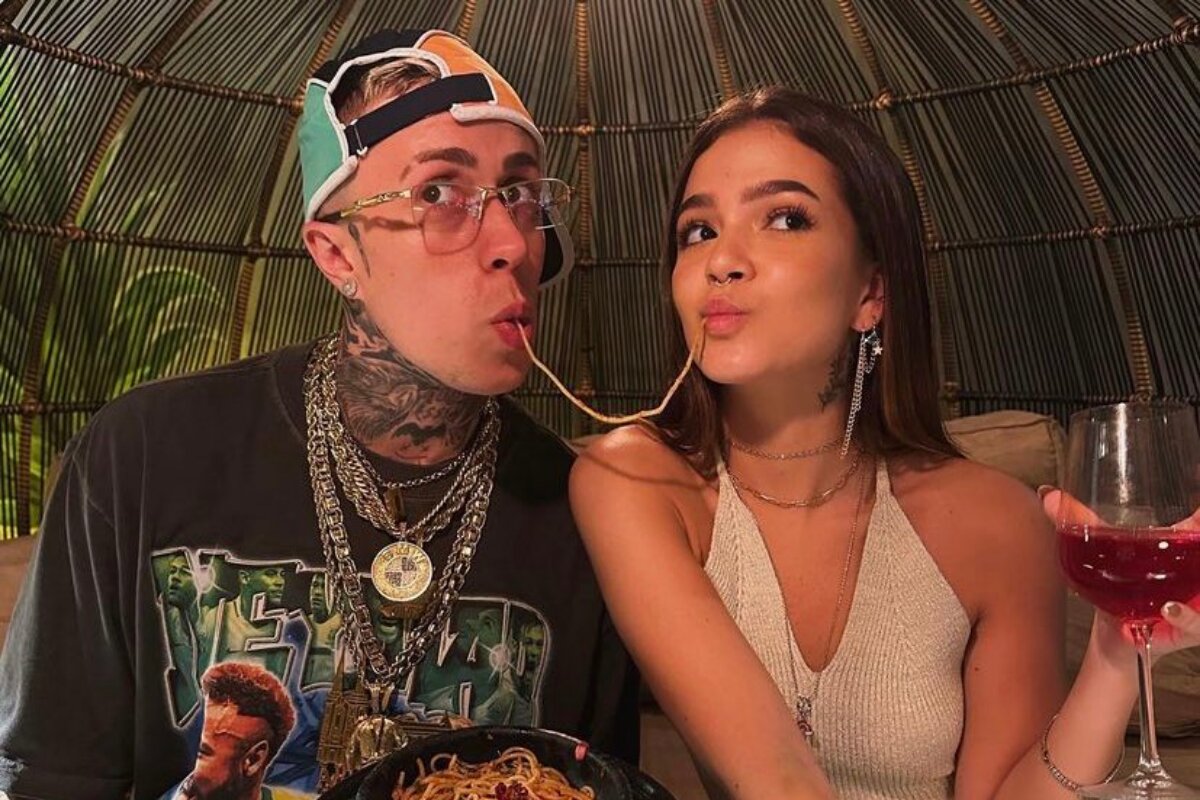 MC Daniel, de boné pra trás,, e Mel Maia, de macacão bege, refazendo a cena clássica do fio de macarrão na boca
