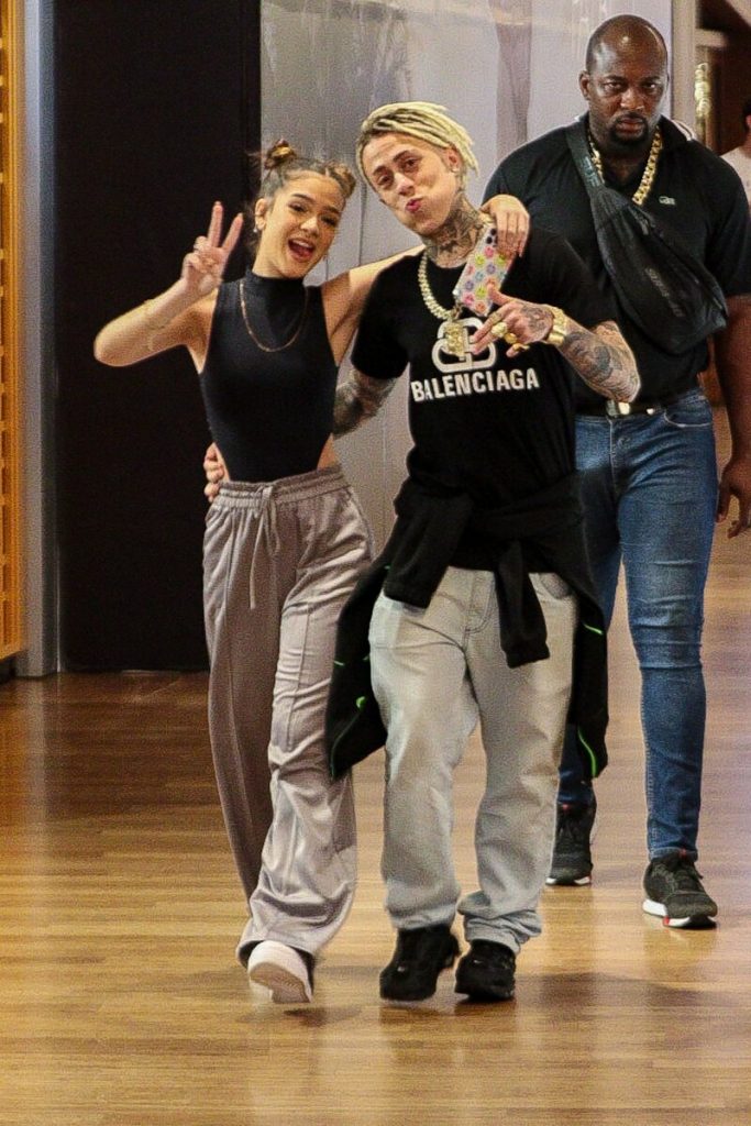 Mel Maia e MC Cabelinho de mãos dadas, escoltados por um segurança, ambos de calça bege e blusa preta