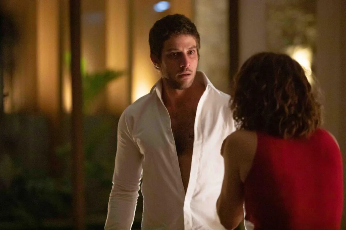 Travessia: Ari (Chay Suede) de camisa social branca discutindo com Núbia (Drica Moraes), de costas, de camisa vermelha