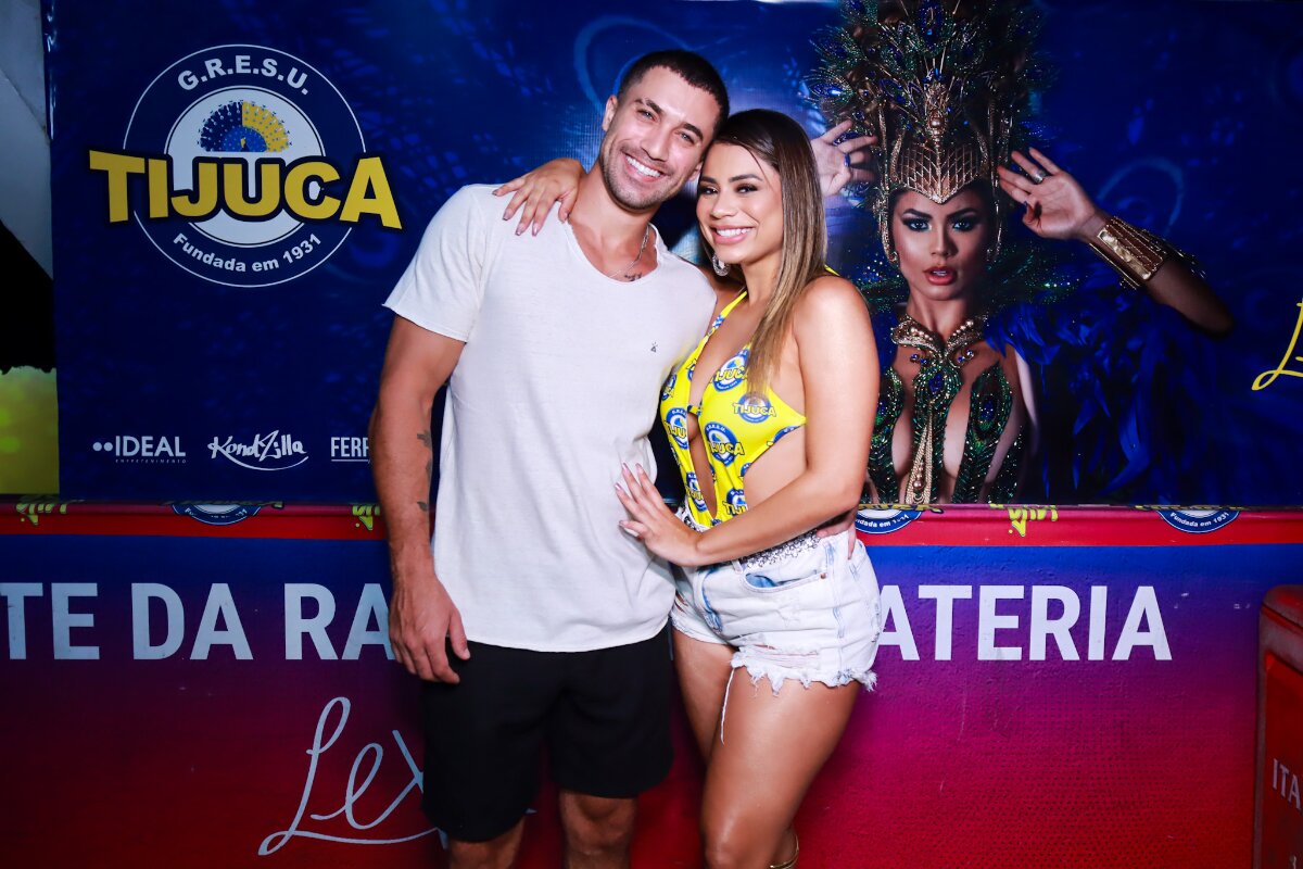 Ricardo Vianna de bermuda preta e camisa básica branca, abraçado a Lexa, que veste short jeans e body de recortes amarelo com detalhes azuis, sandálias de salto com tiras até o joelho