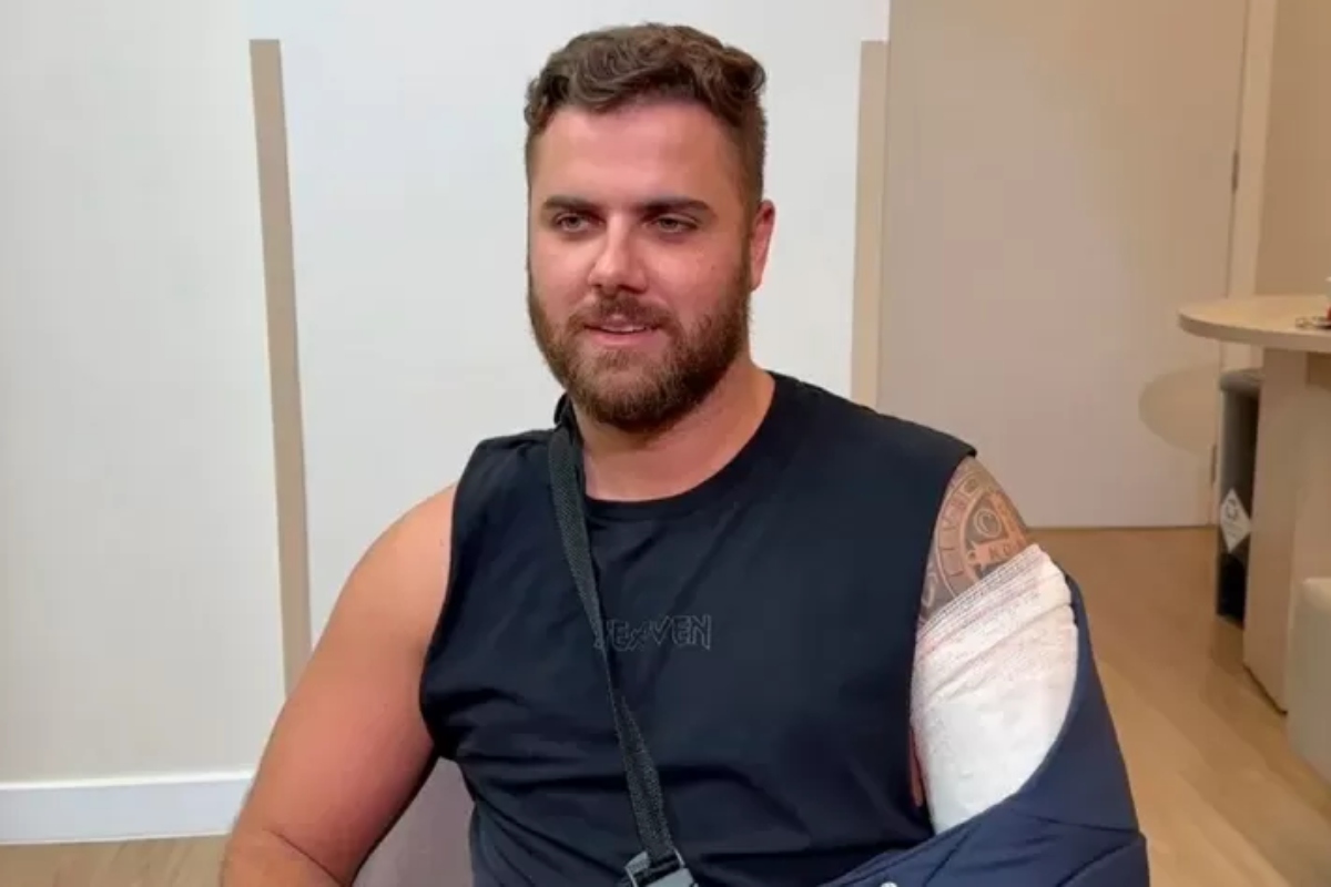 Zé Neto sentado, de camisa preta regata, com braço imobilizado