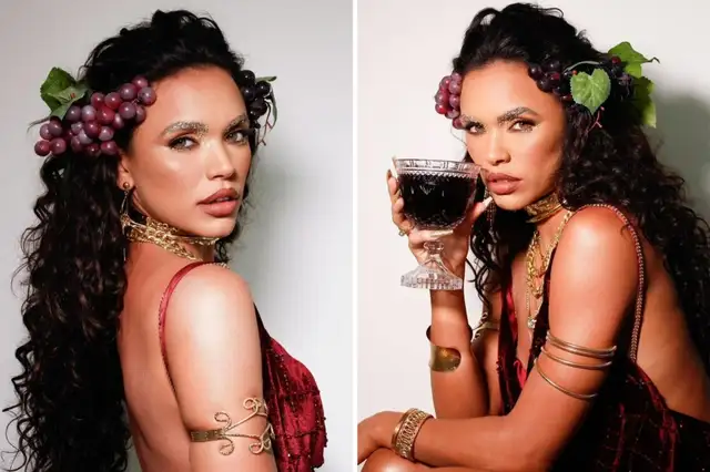 Giovana Cordeiro de vestido cor de vinho, segurando uma taça, com adereços de uva nos cabelos